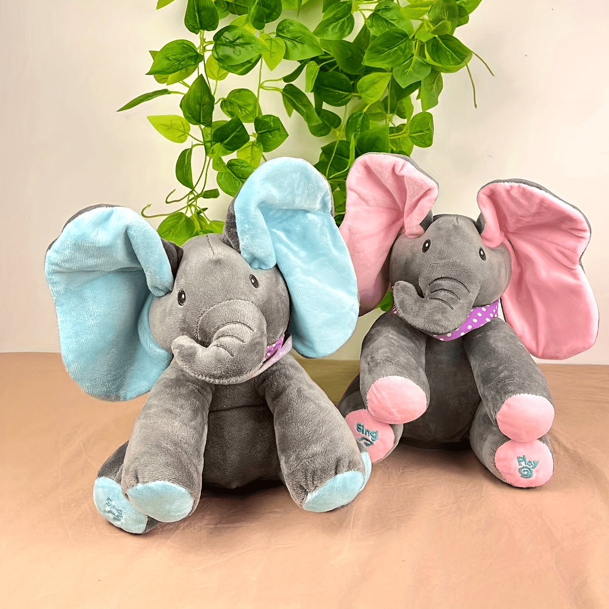 Two plush elephants with blue and pink ears on a beige surface with green leaves in the background