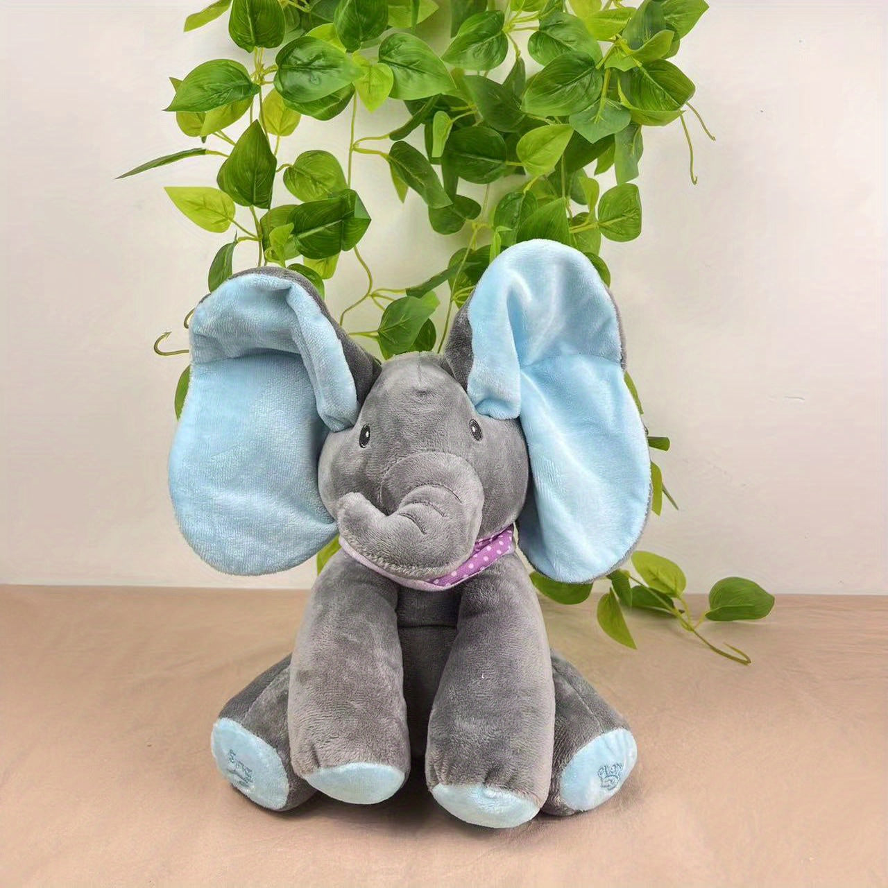 Plush elephant toy with blue ears and feet on a beige surface with green leaves in the background