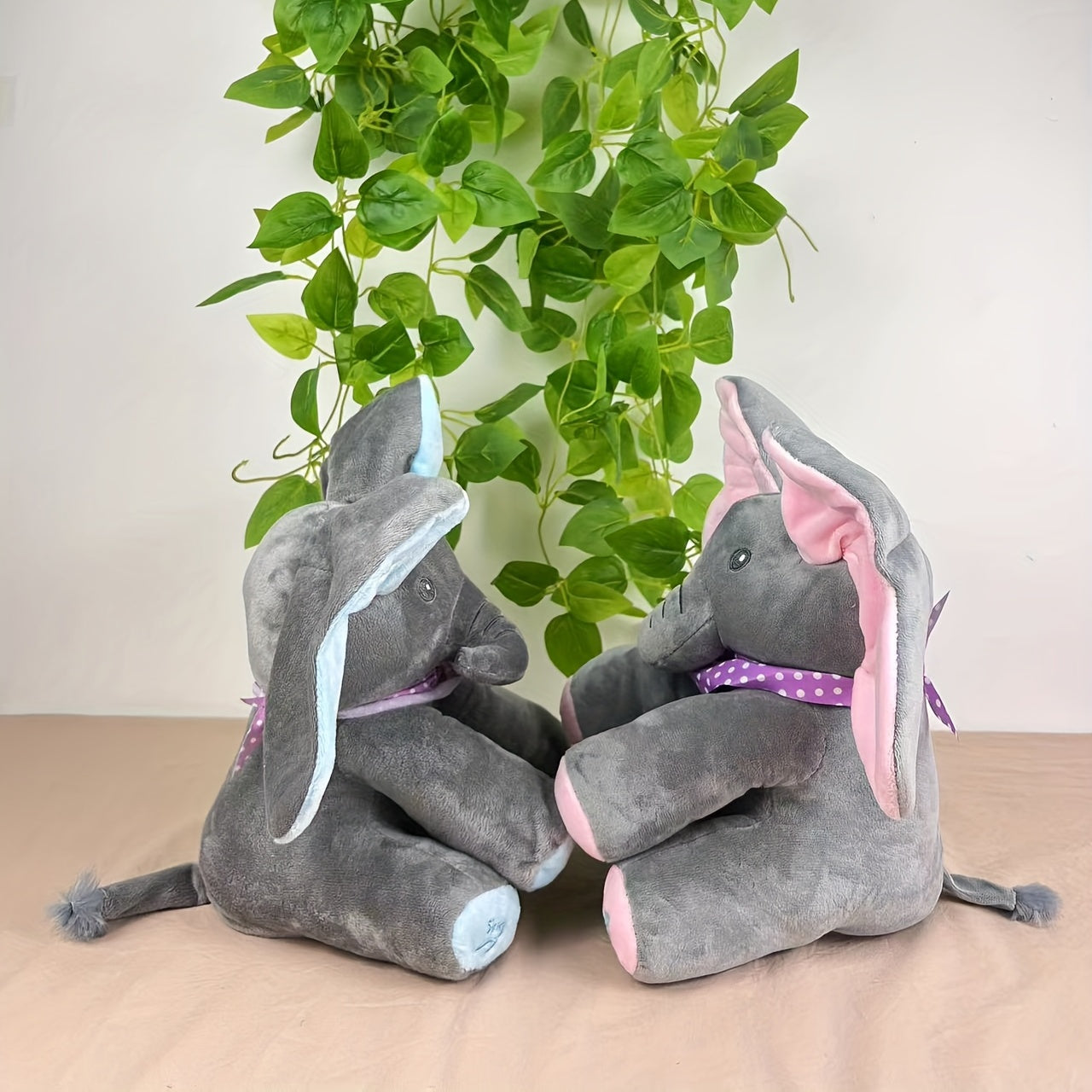 Two gray elephant plush toys with colorful accents sitting on a beige surface with green leaves in the background.