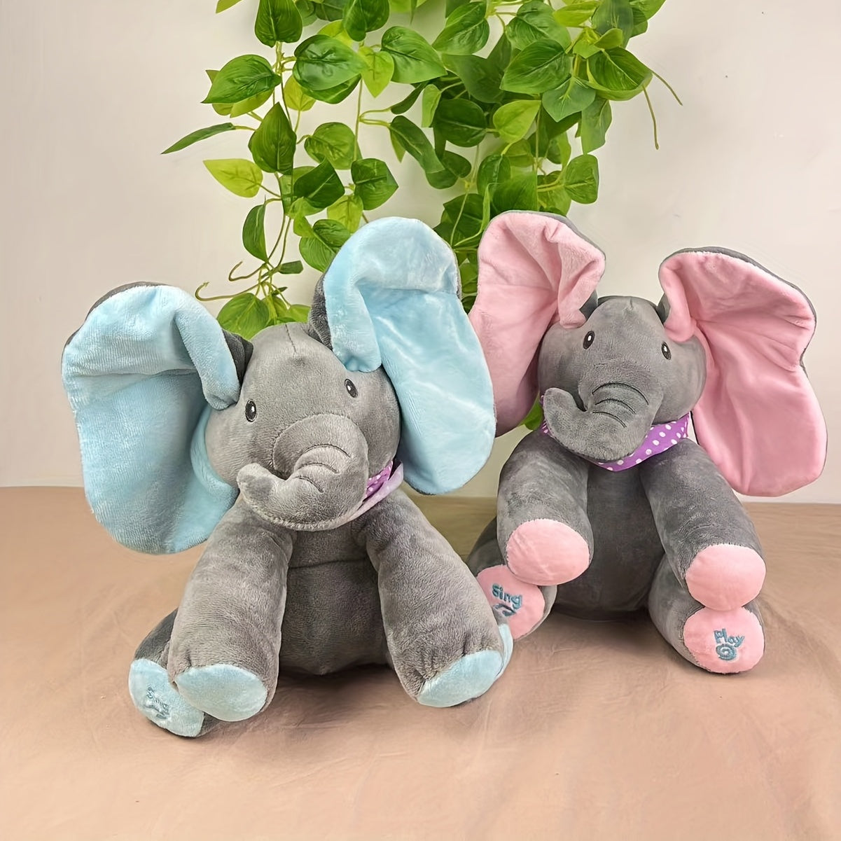 Two plush elephants, one with blue ears and the other with pink ears, sitting on a wooden surface with a plant in the background.
